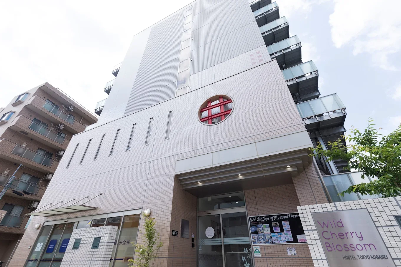 A modern multi-story hostel building with a tiled facade, glass balconies, and a circular window with a red grid design, located on a city street in Tokyo, Japan.