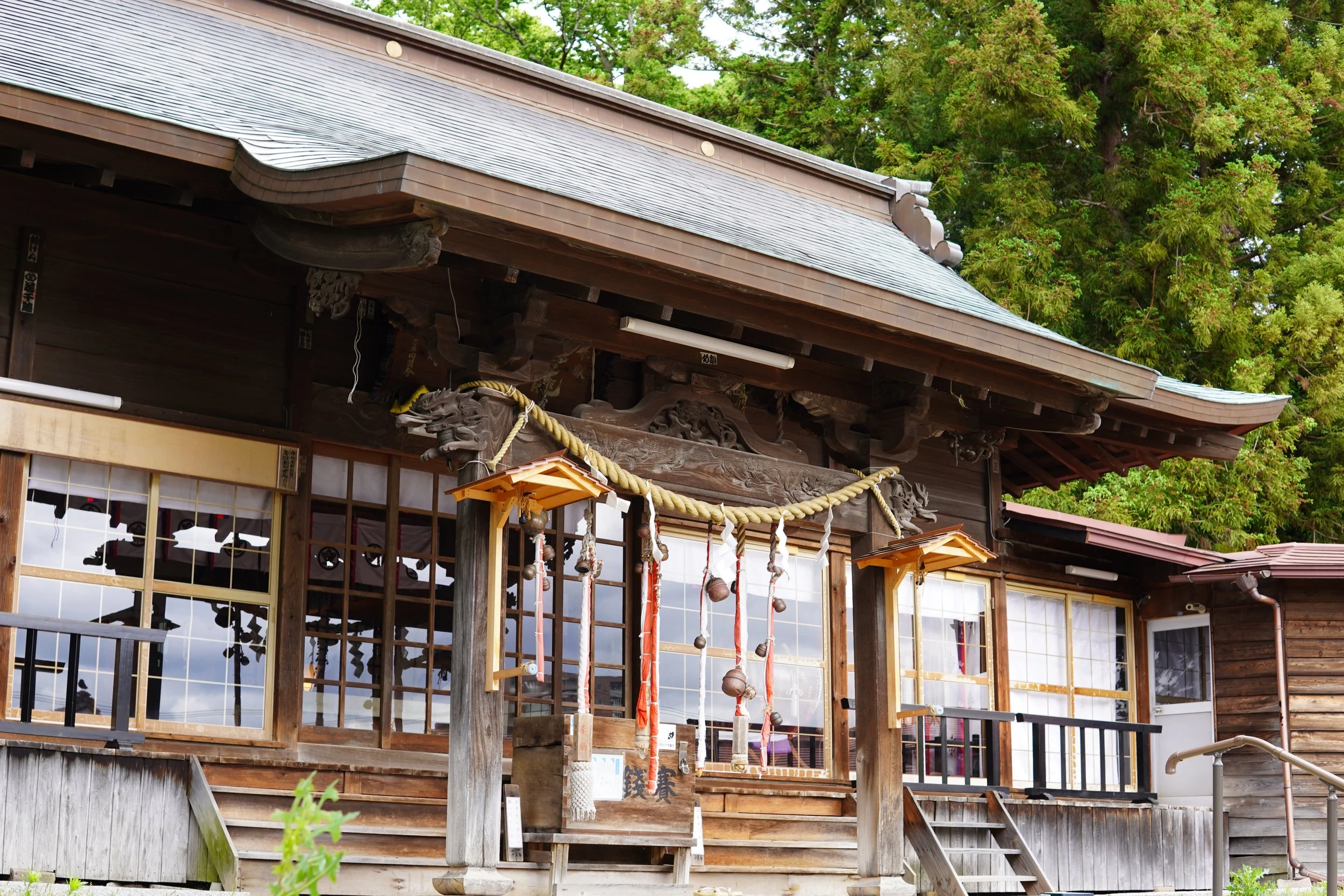 木造の神社の建物で、竹や木の飾りや鈴が掛けられている。背景には緑の木々が茂っている。