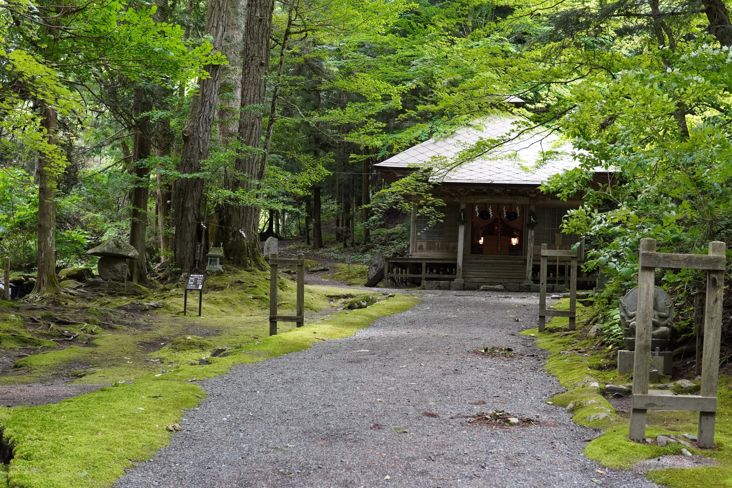 緑豊かな森の中に伝統的な木造の建物があり、周囲に石像や木製のしめ縄が設置されている。
