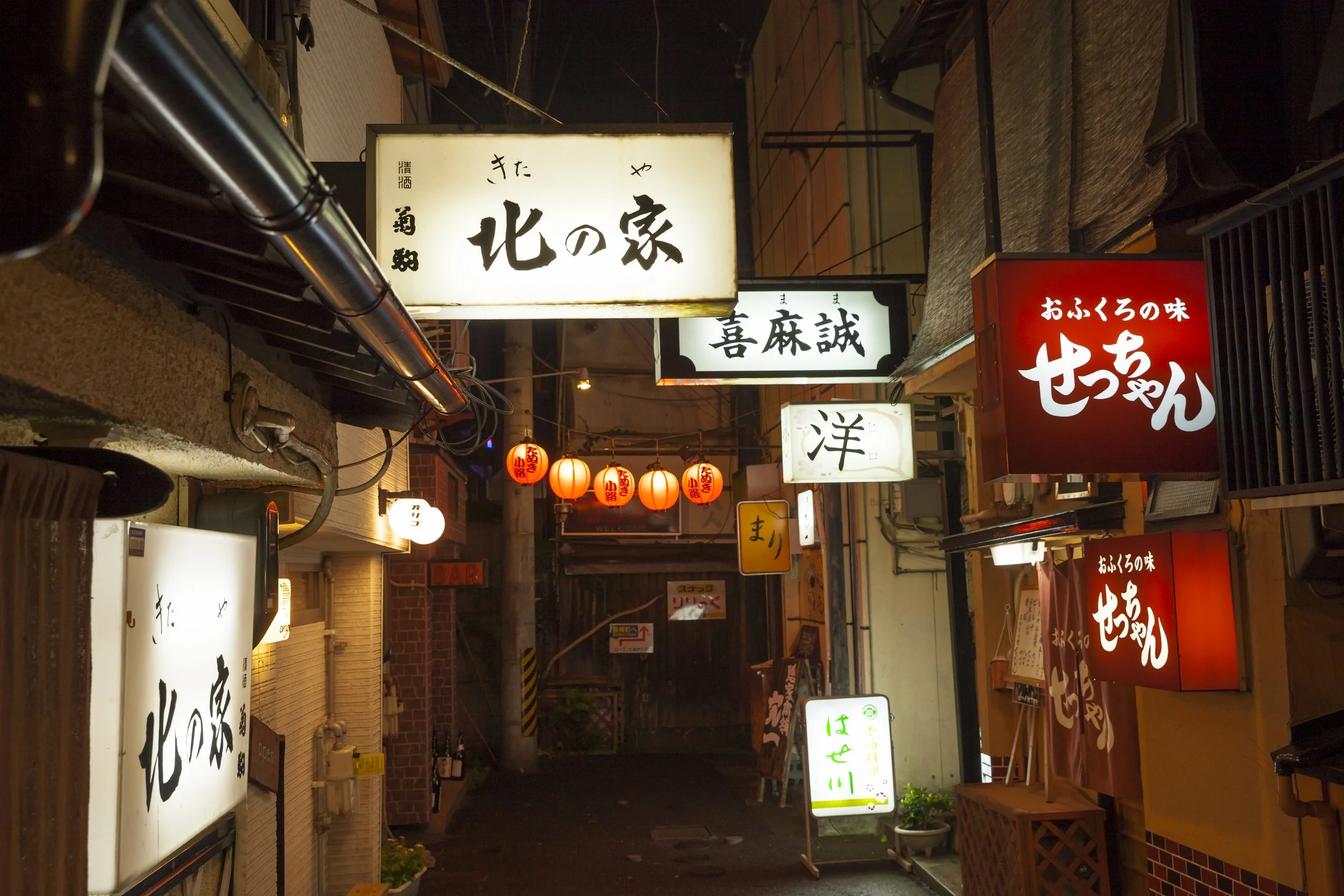 Nighttime street scene with illuminated Japanese restaurant signs and red lanterns hanging overhead.