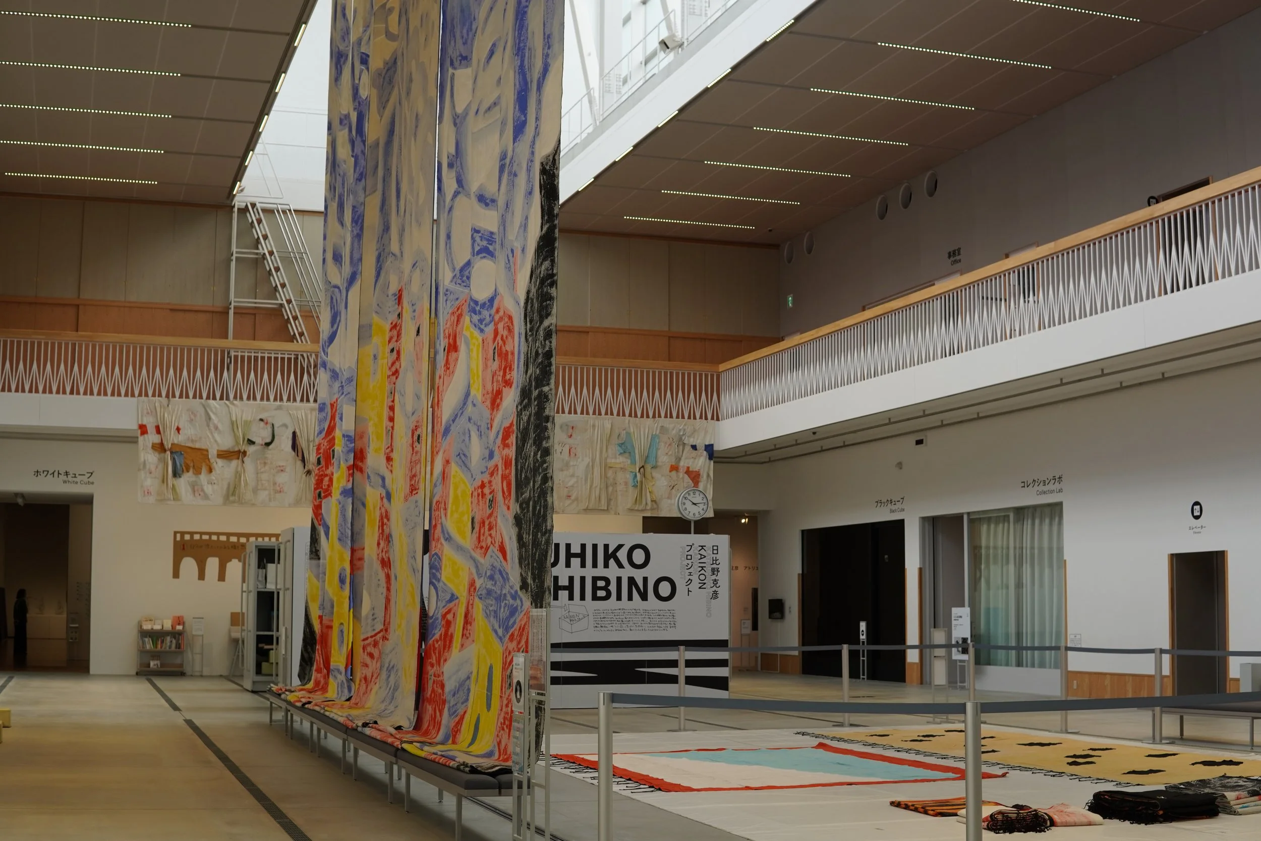 美術館やギャラリーの展示スペースに、色とりどりのテキスタイルアートや壁掛け作品が展示されている。大型の布作品が垂れ下がり、その他の作品も床に広げられている。高い天井と木製の壁枠、照明に照らされた展示空間。