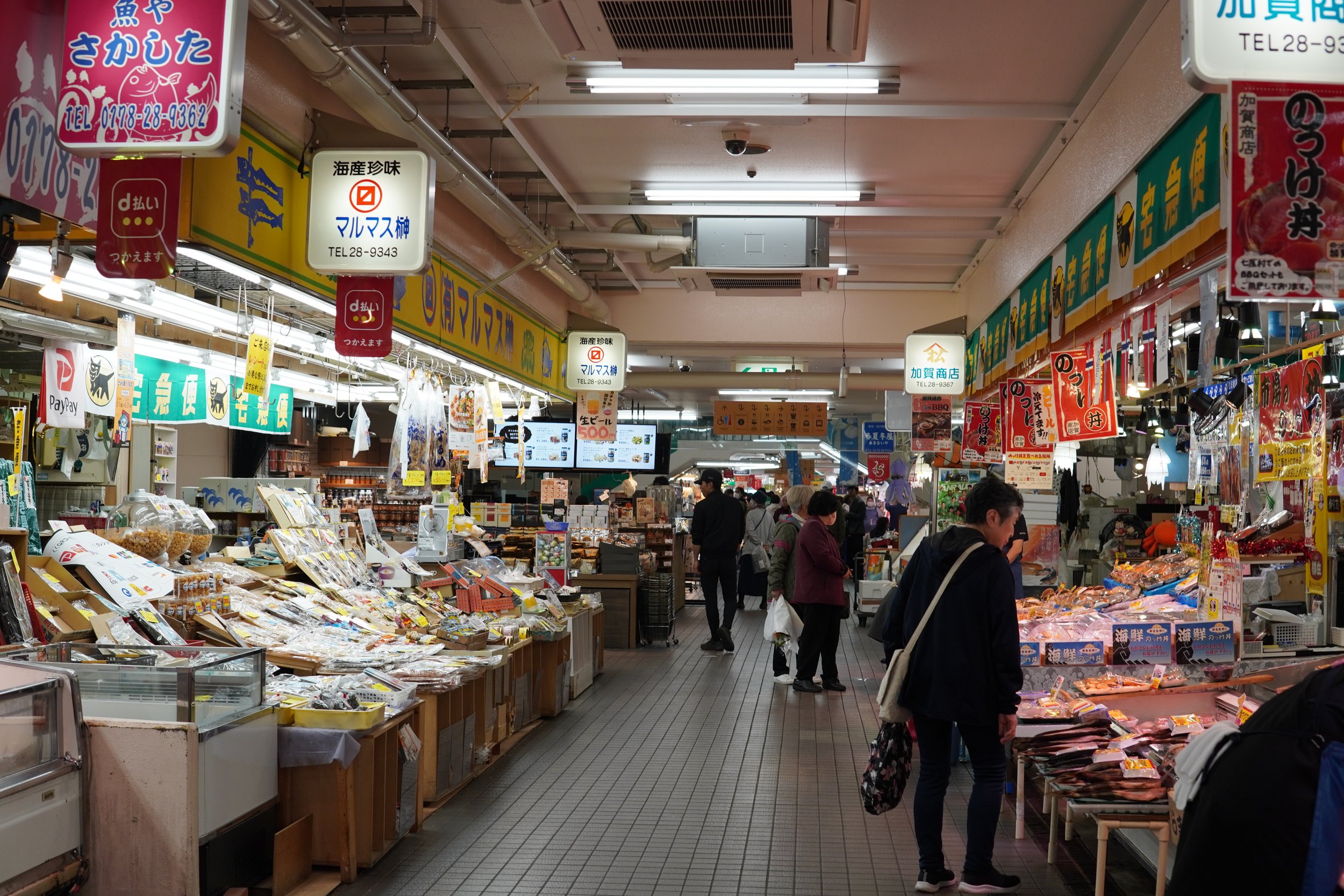 店内の市場の様子で、多くの人が魚や肉などの食品を見ている。棚にはさまざまな商品が並んでいる。