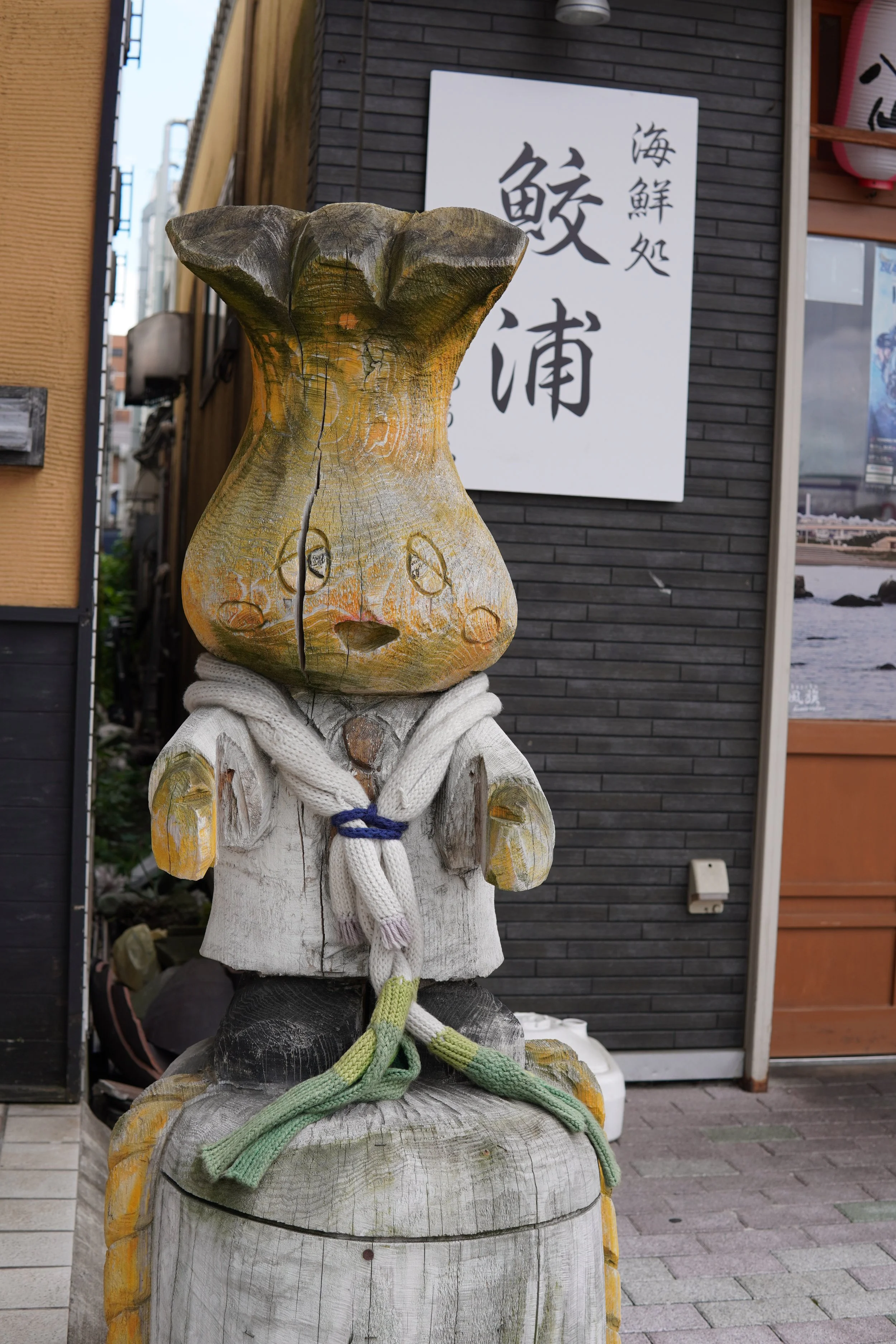 木彫りのウサギの像で、その頭は木の割れ目によって顔の形になっている。人形には白いスカーフと緑のロープが巻かれている。背景には看板と建物がある。