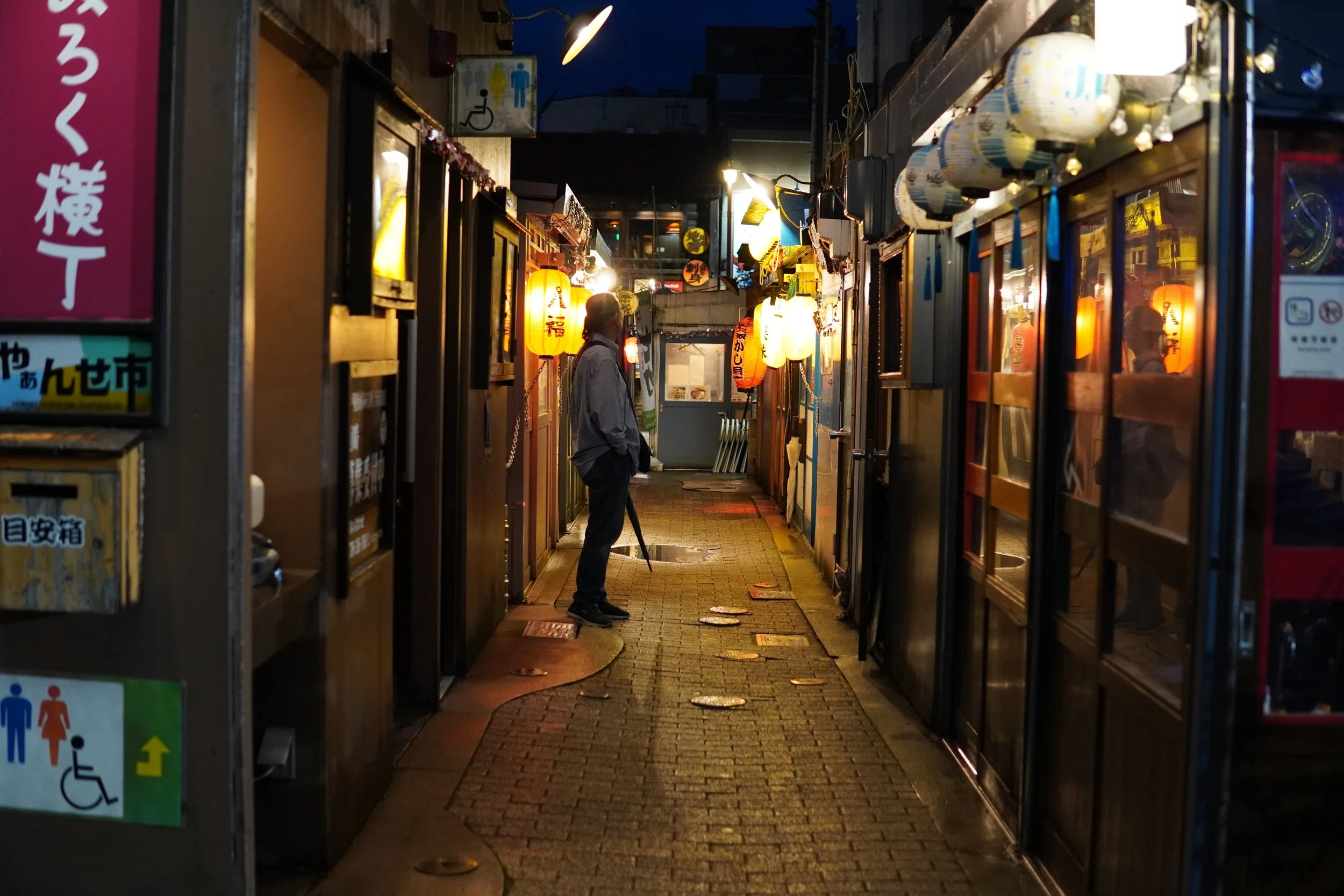 夜の小さな飲食店の通路に、提灯や電飾のライトが灯る。男性が傘を持ち立っている。