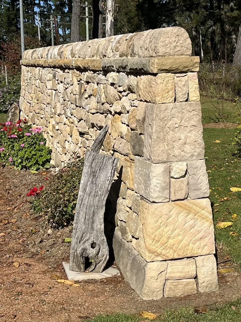 A stone wall constructed with large, irregularly shaped rocks, with a piece of weathered driftwood leaning against it, in a garden with flowers and greenery in the background.