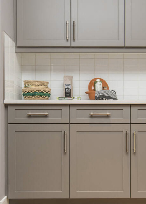 Kitchen countertop with woven baskets, a black box, a brown dish with a white bottle, and black and white napkins. Grey cabinets with silver handles above and below.