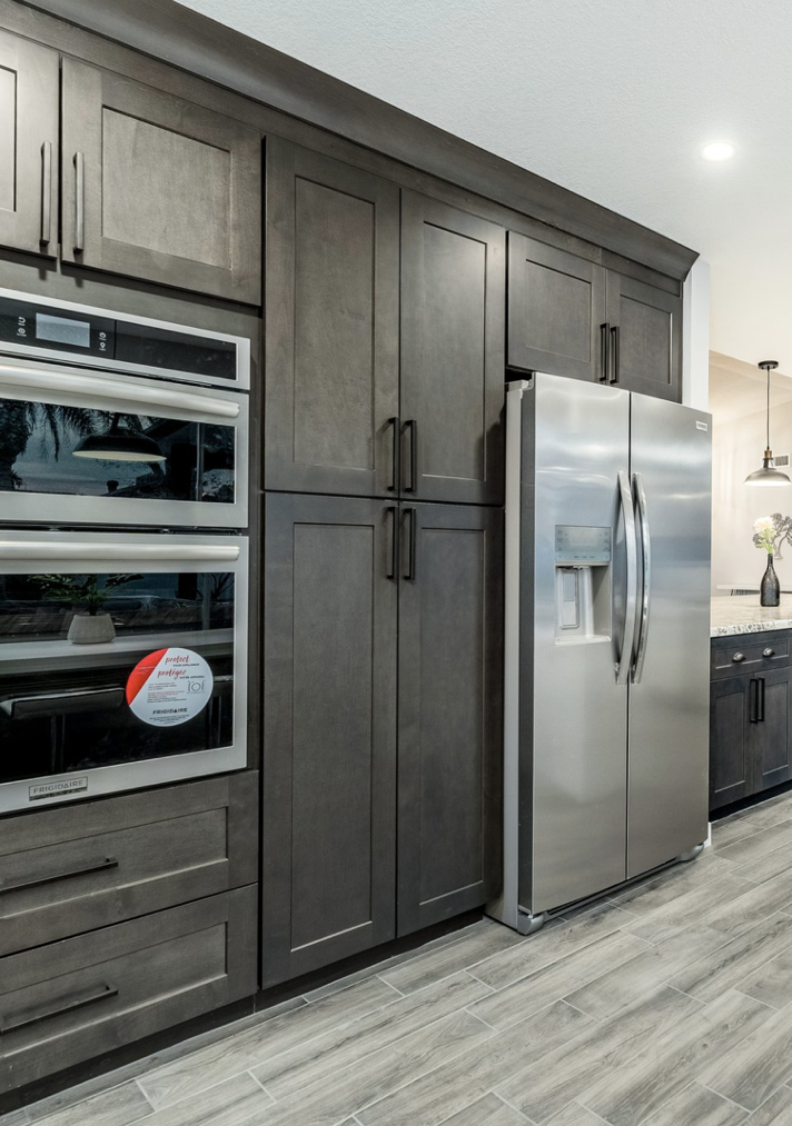 Modern kitchen with dark wood cabinets, stainless steel double oven, and side-by-side refrigerator with water and ice dispenser, on a gray tiled floor.
