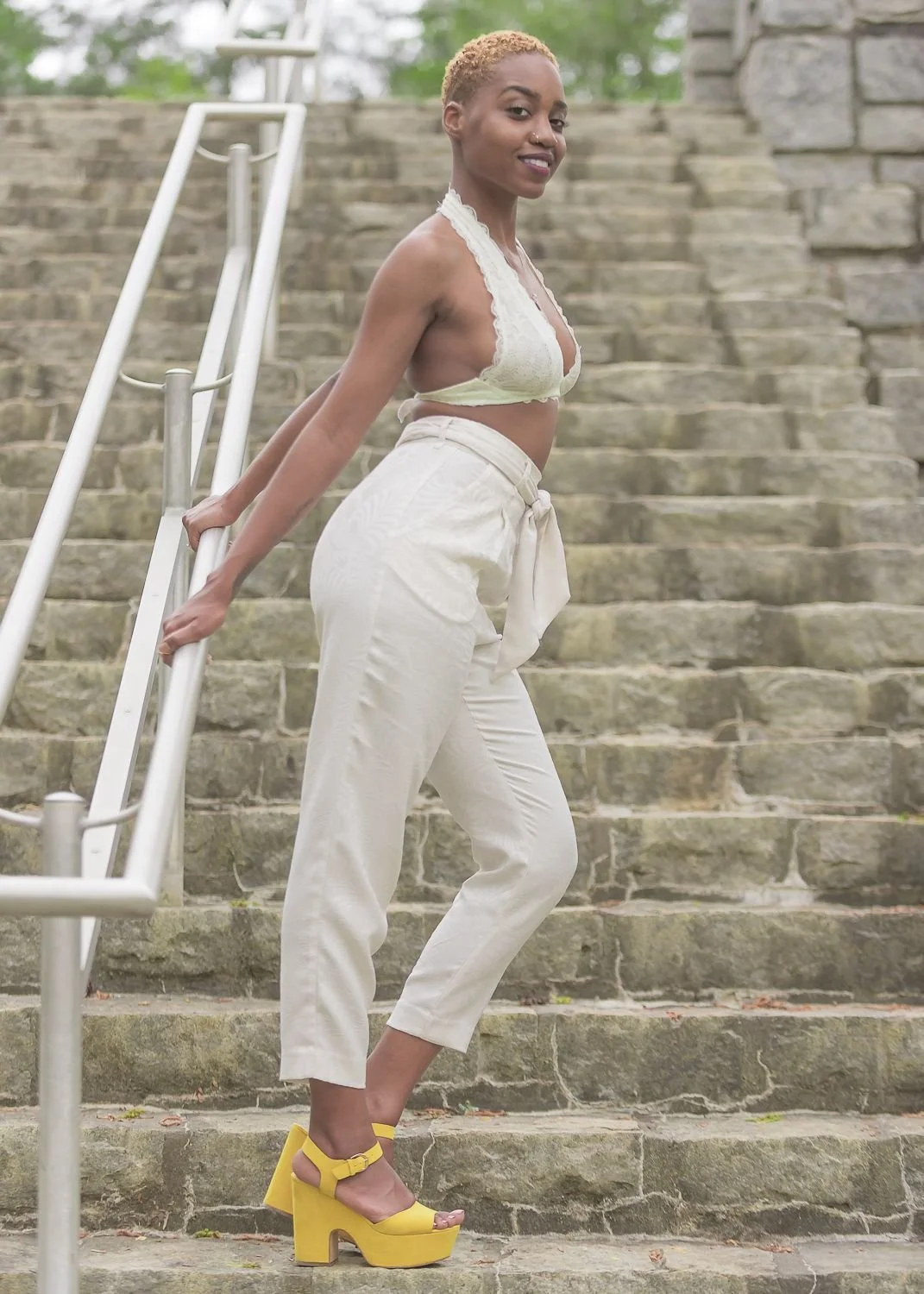 A woman with short, blonde hair, wearing a light yellow lace crop top, high-waisted cream-colored pants tied at the waist, and bright yellow high-heeled sandals, stands on stone stairs outdoors, holding onto a metal handrail and looking at the camera