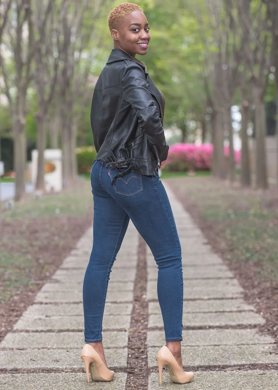 A woman with short blonde hair, wearing a black leather jacket, dark blue jeans, and beige high heels, standing on a park pathway lined with trees and pink flowering bushes, smiling while looking over her shoulder.