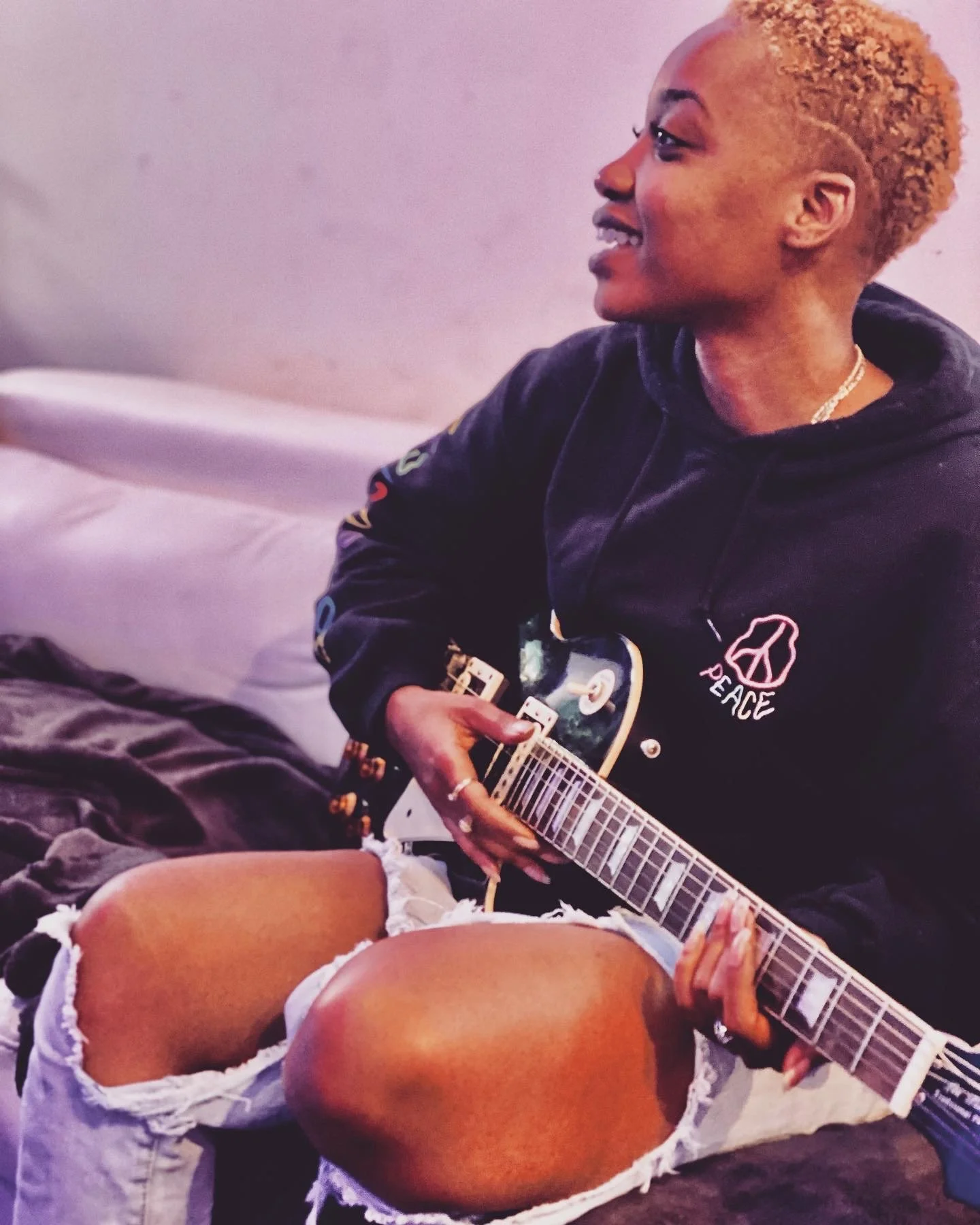 Young woman with bleached blonde hair playing an electric guitar. She is sitting on a couch wearing a black hoodie with a peace sign and the word "peace" on it. She is also wearing ripped white denim shorts.