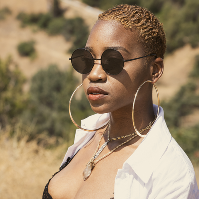 Woman with short blonde hair wearing round black sunglasses and large gold hoop earrings, posing outdoors with a desert landscape in the background.