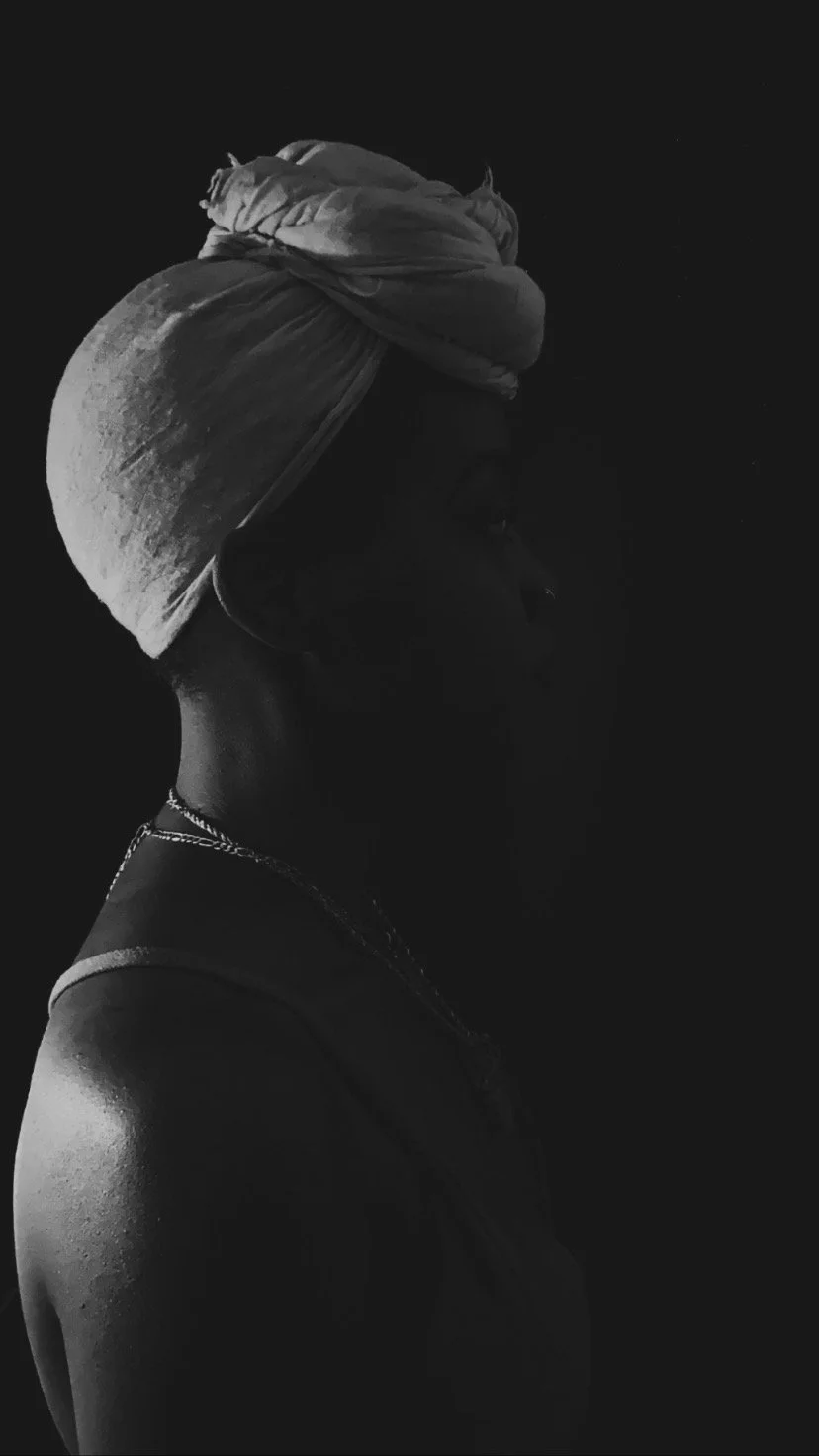 Side profile of a woman with a headwrap, jewelry, and sleeveless top, in black and white.