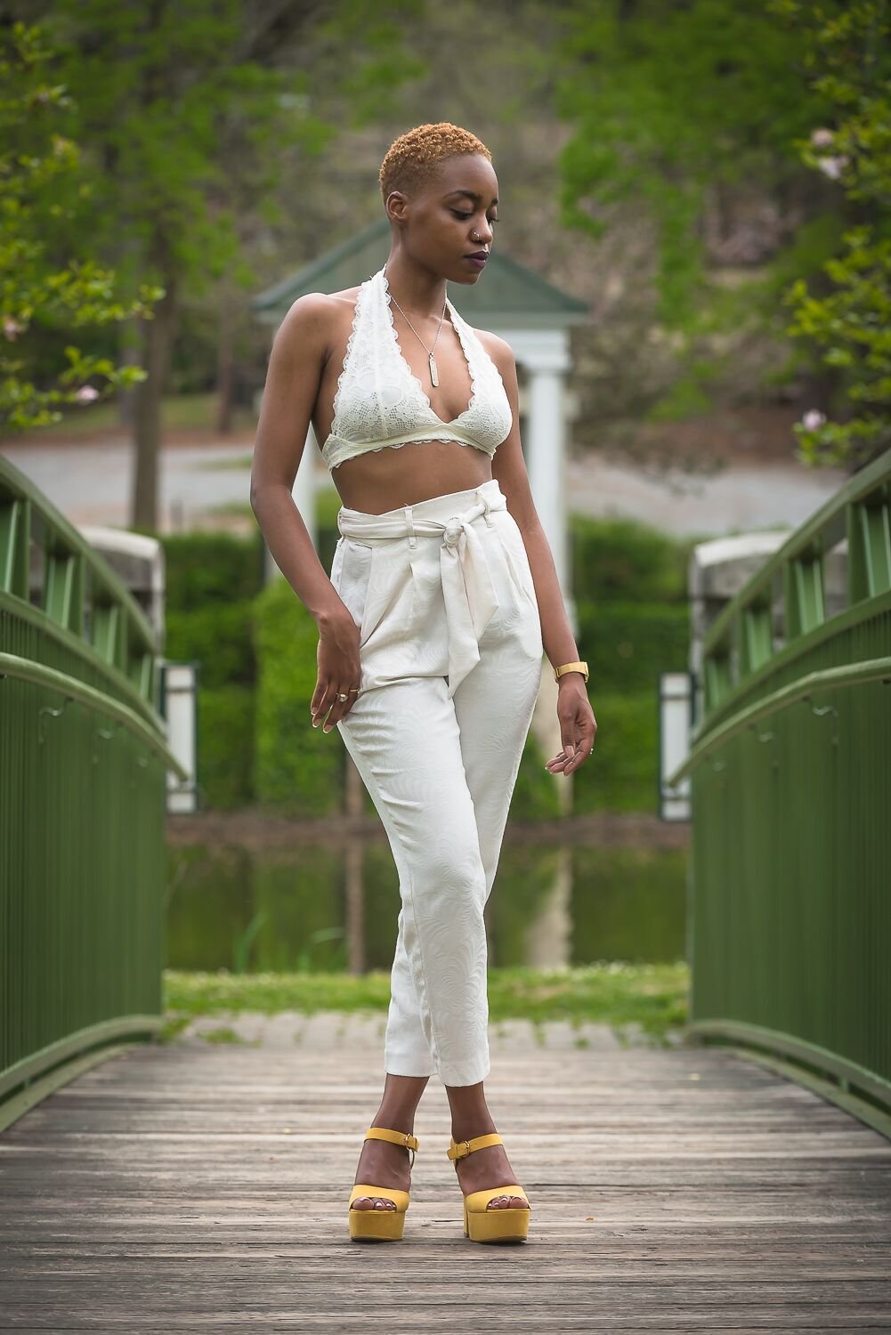 A woman with short, curly hair wearing a white lace crop top, high-waisted white pants, and yellow platform sandals standing on a wooden bridge in a park with green trees and water in the background.