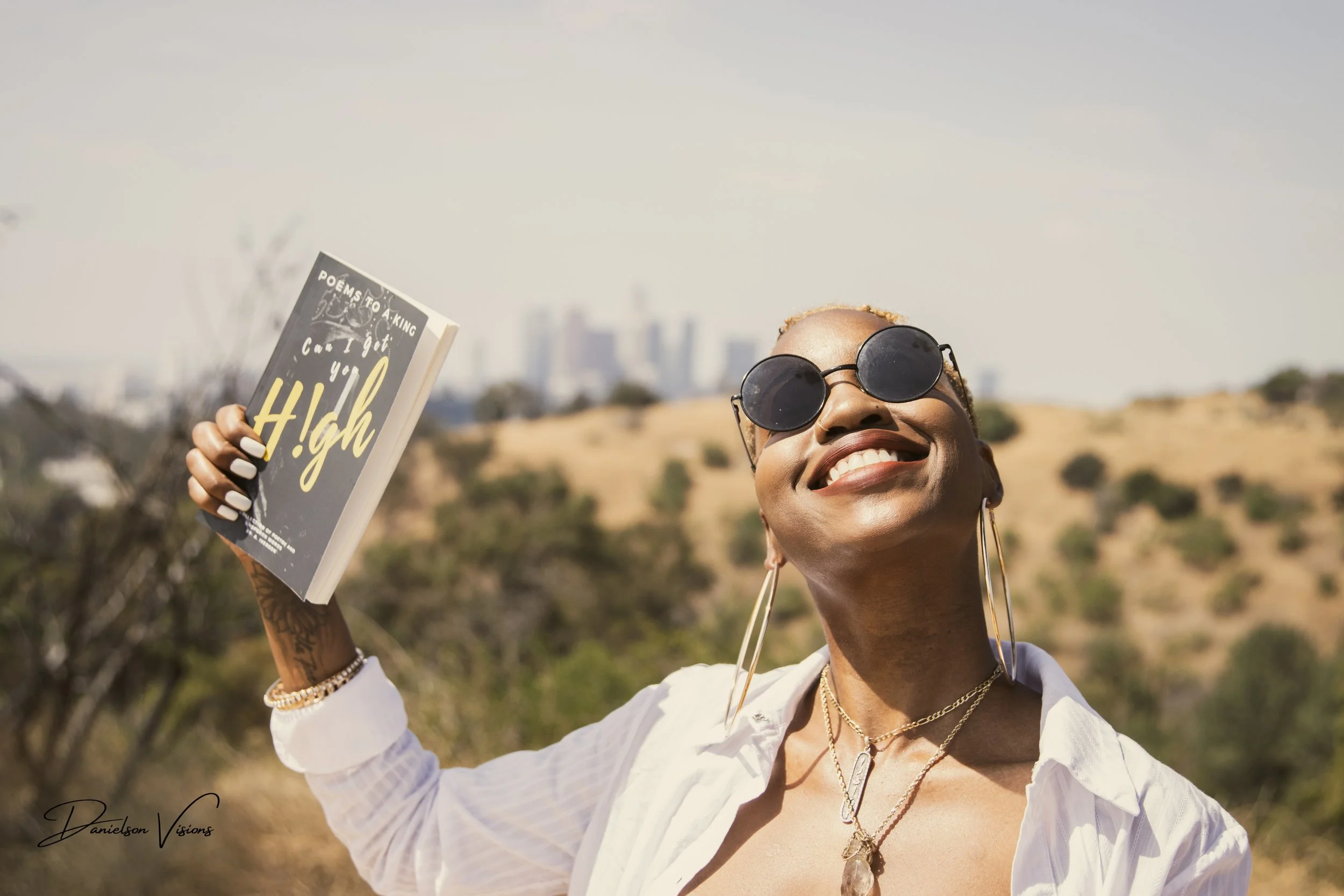 A smiling woman with short blond hair wearing sunglasses, a white shirt, and gold jewelry, holding a book titled 'Poems to a King' outdoors with a blurred background of trees and hills.