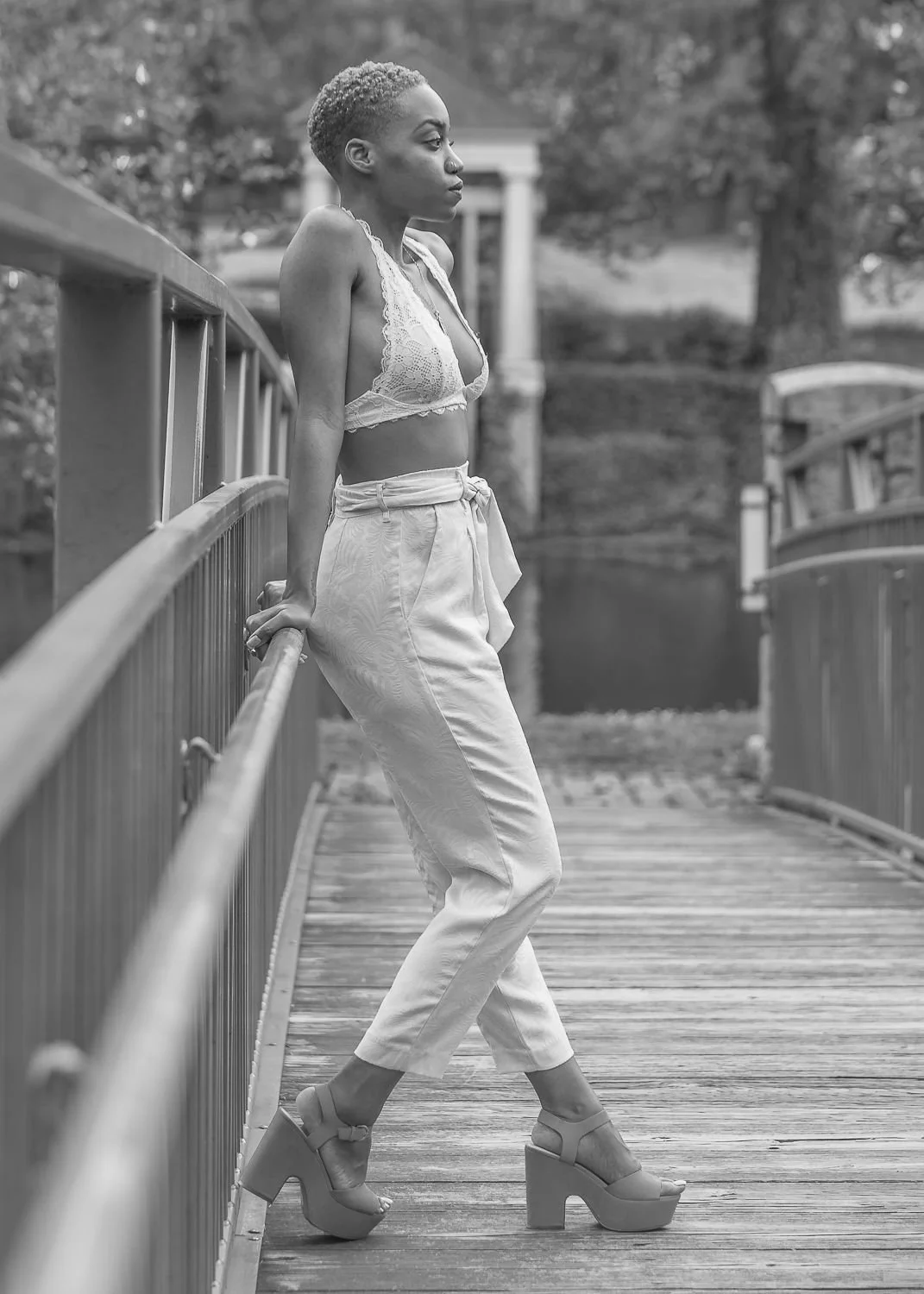 A woman with short hair standing on a wooden bridge, leaning against the railing, looking to the right, wearing a lace crop top, high-waisted pants, and platform heels.