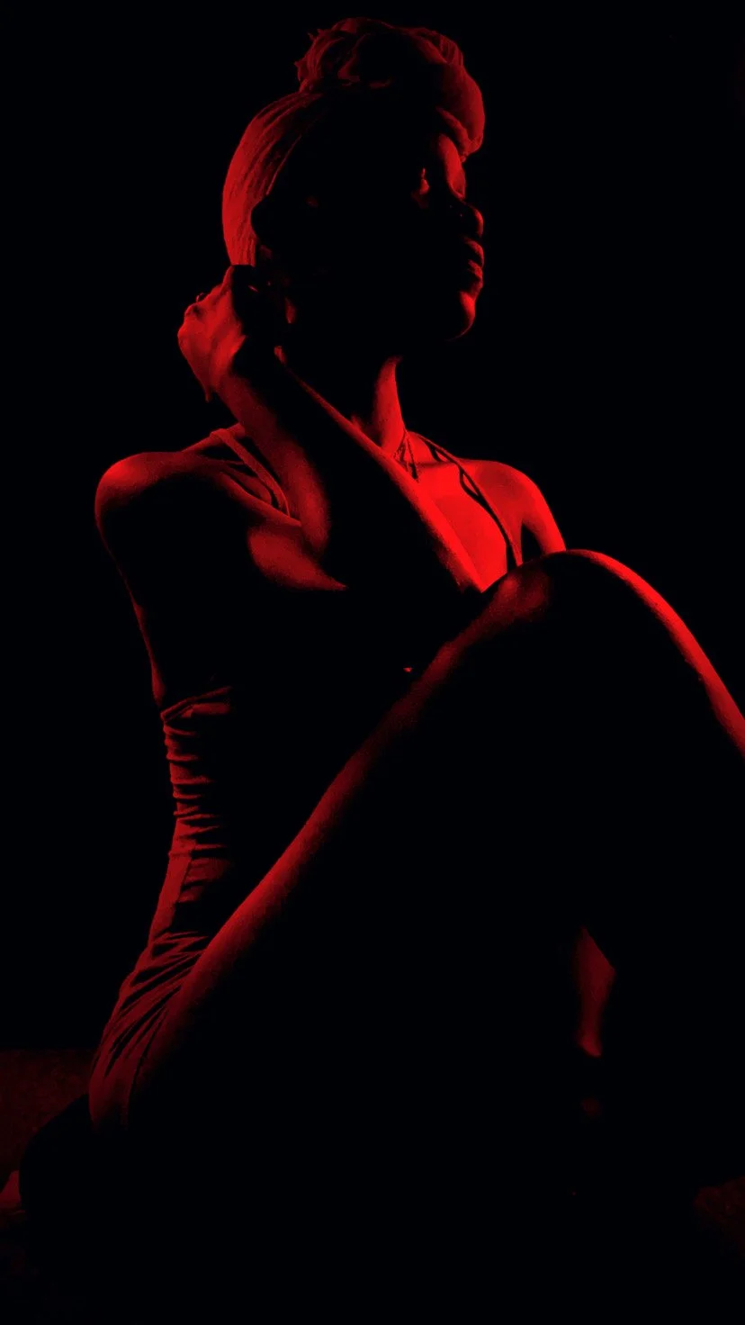 Silhouette of a woman with short hair, sitting with one knee up, illuminated by red light against a dark background.