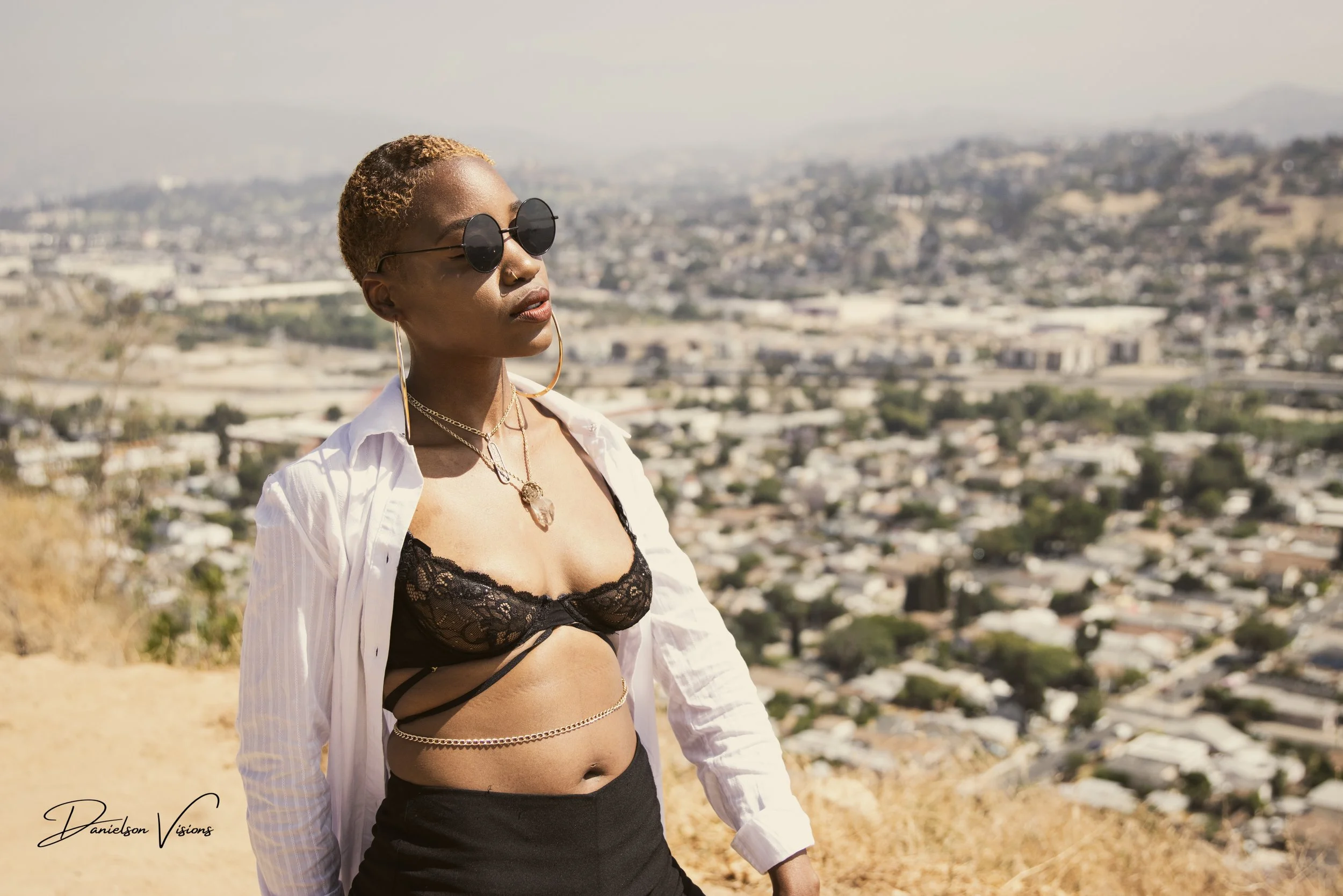 Woman with short blonde hair wearing sunglasses, a black lace bralette, a white open shirt, and layered jewelry, standing outdoors with a cityscape and hills in the background.