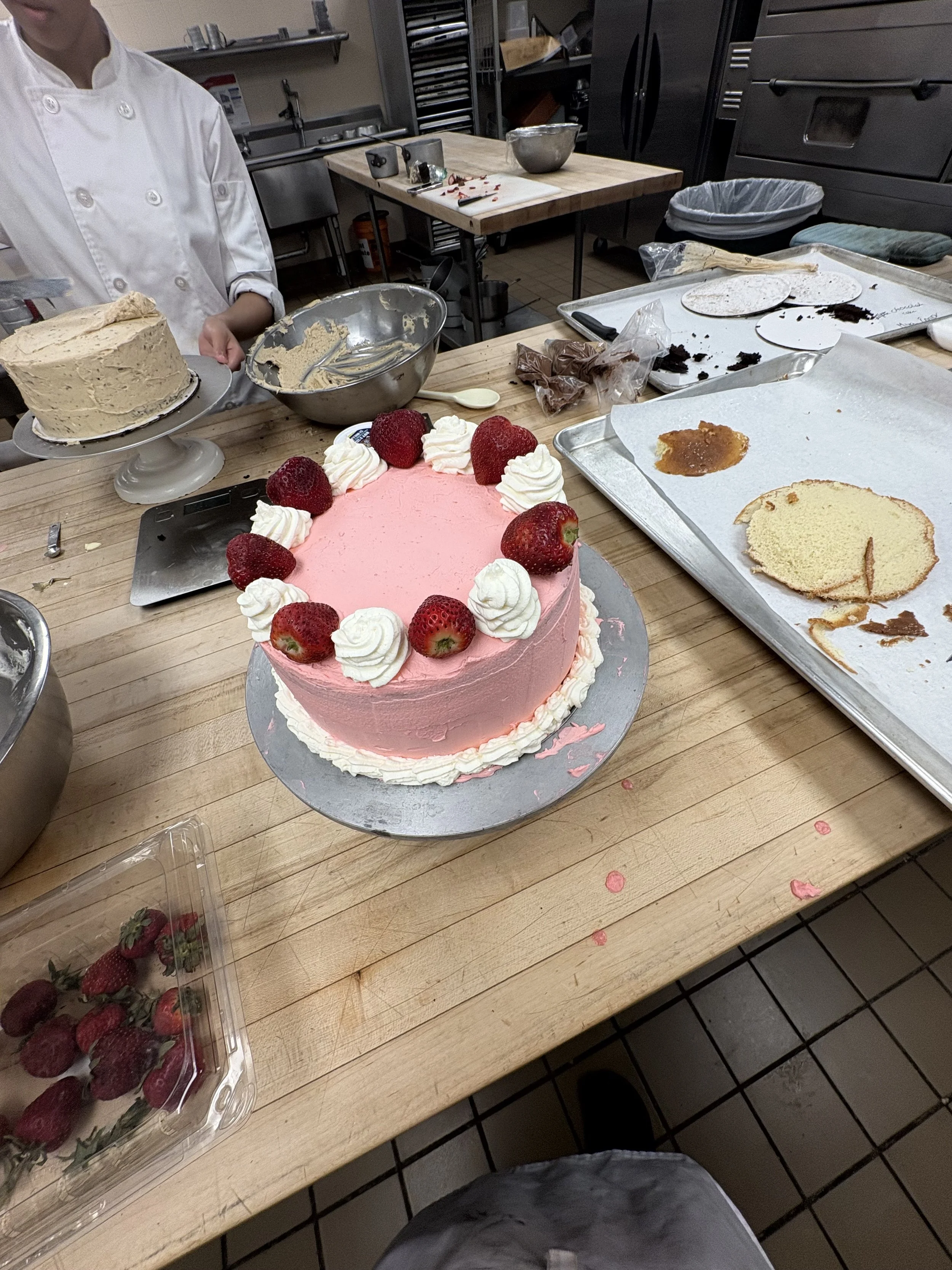 A pink frosted cake decorated with whipped cream and fresh strawberries on top, with a second layer of cake partially cut and in the background. There are various baking tools and ingredients on the baking table in a commercial kitchen.
