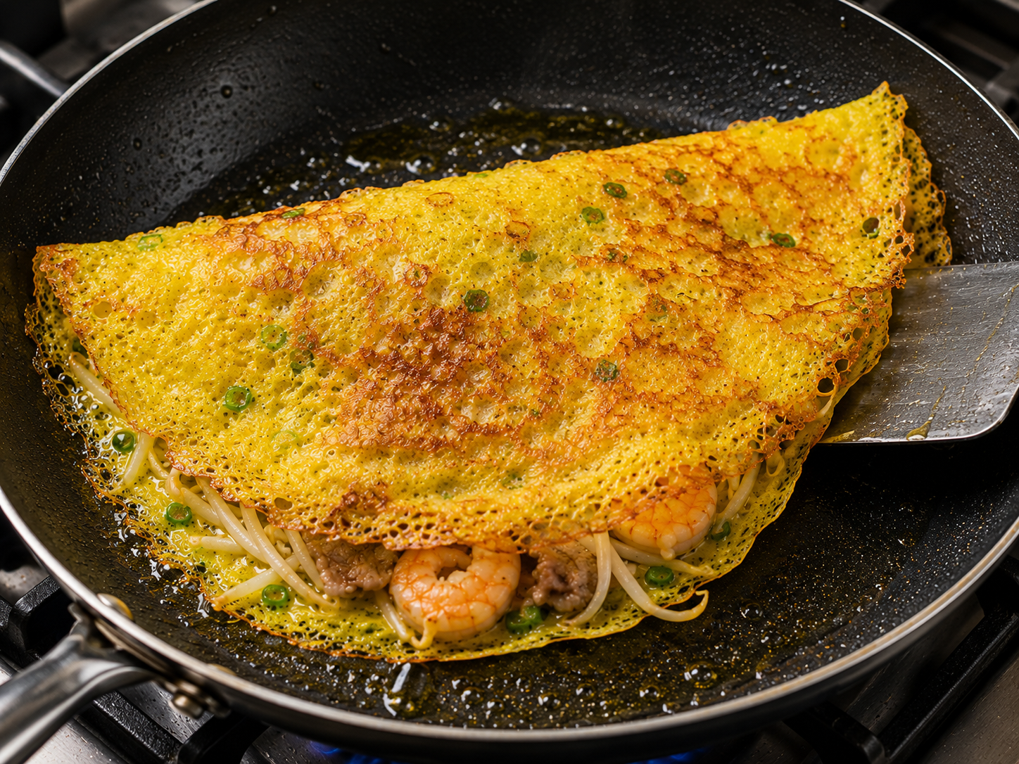 A golden bánh xèo pancake crisping in a hot skillet with lacy edges, shrimp, pork, bean sprouts, and a spatula lifting the edge.