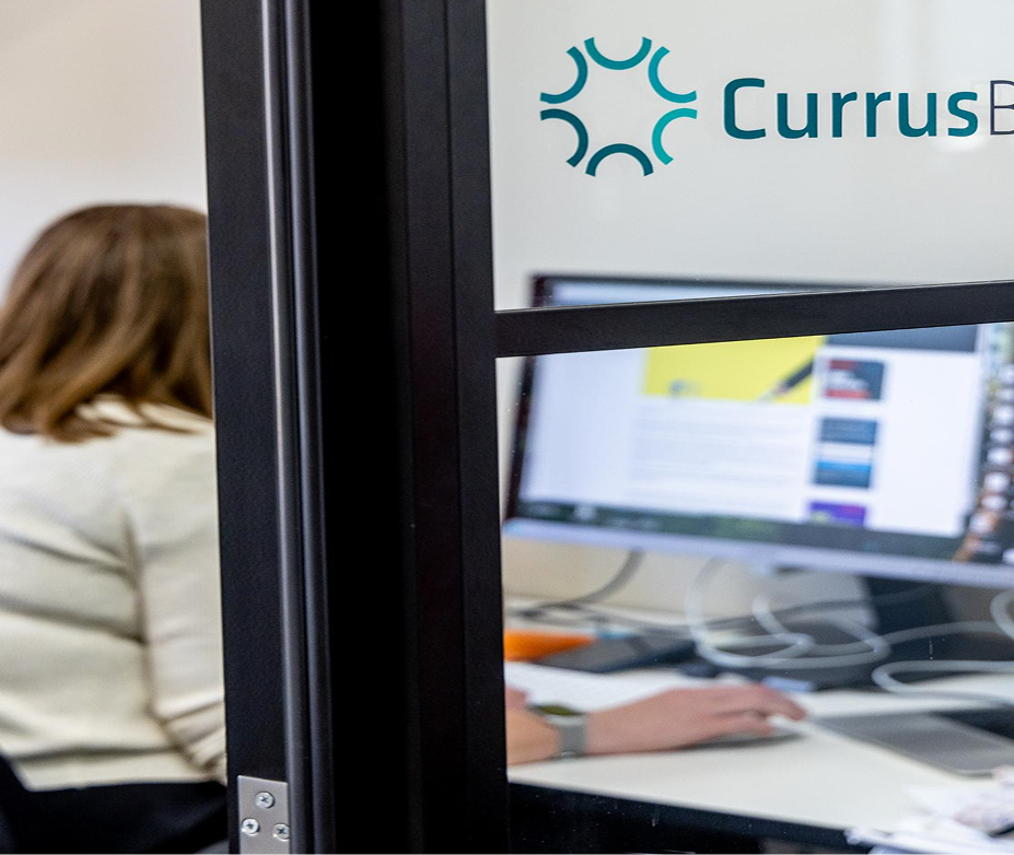 A person working at a computer in an office cubicle with a sign that reads 'Currus' and a logo.