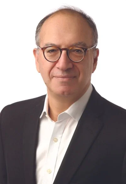 A middle-aged man with glasses, wearing a black suit and white shirt, posing against a white background.