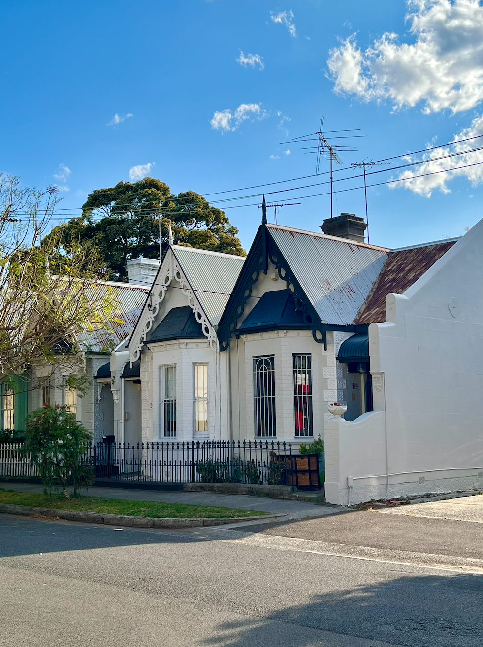 A white Victorian-style house with ornamental trim and barred windows, set on a street corner under a blue sky with clouds.