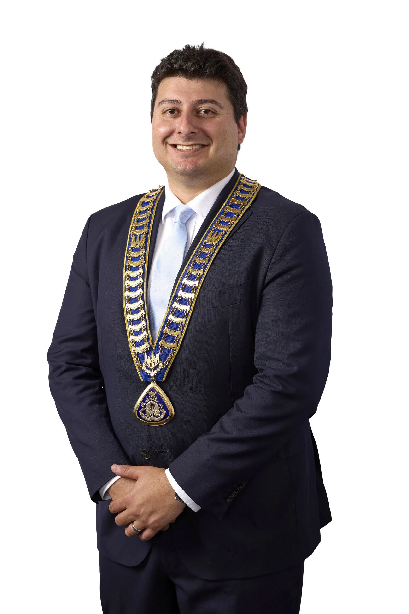 A man in a dark suit and light blue tie wearing Masonic regalia with blue and gold sash and pendant, standing against a plain white background.