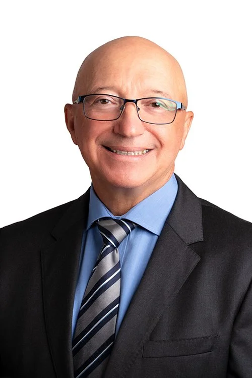 A bald man wearing glasses, a black suit, blue shirt, and a striped tie, smiling against a white background.