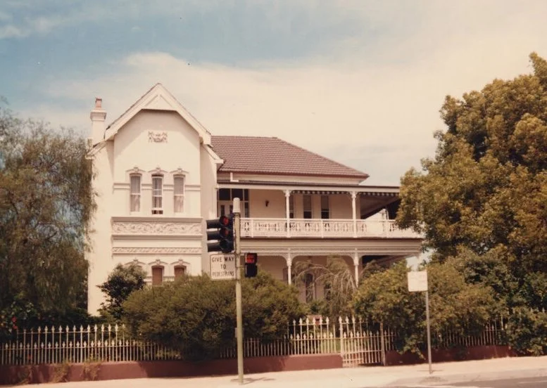 A large, Victorian-style house with a white exterior and a wraparound porch, surrounded by trees and bushes, in a residential neighborhood.