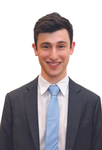 A young man smiling, dressed in a dark suit with a white shirt and a light blue tie.