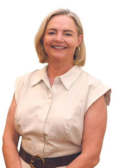 A smiling middle-aged woman with blonde hair, wearing a beige sleeveless blouse and a black belt, standing against a white background.