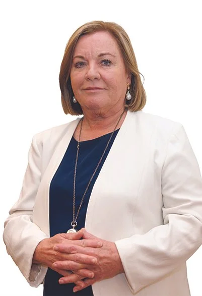 A woman with shoulder-length light brown hair, wearing a white blazer over a navy blue top, and jewelry including earrings and a long necklace, standing against a plain white background.