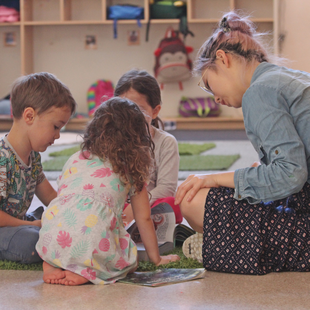 A group of four children sitting on a carpeted floor, closely gathered, looking at a book or magazine together in a playroom or classroom setting.