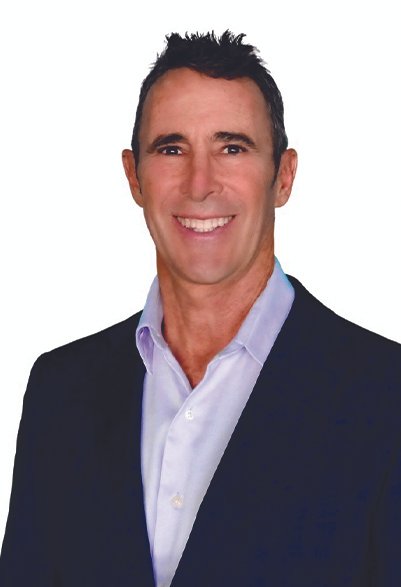 A smiling man with short dark hair wearing a dark suit jacket and a light blue dress shirt against a white background.