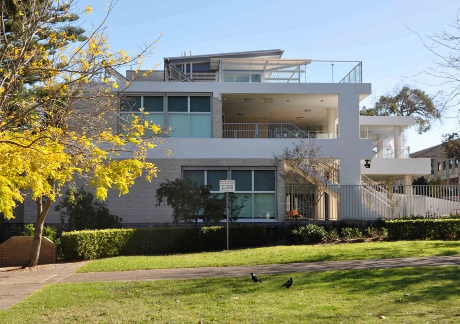 Modern multi-story apartment building with balconies, surrounded by green lawn, trees, and birds in a park-like setting on a clear day.