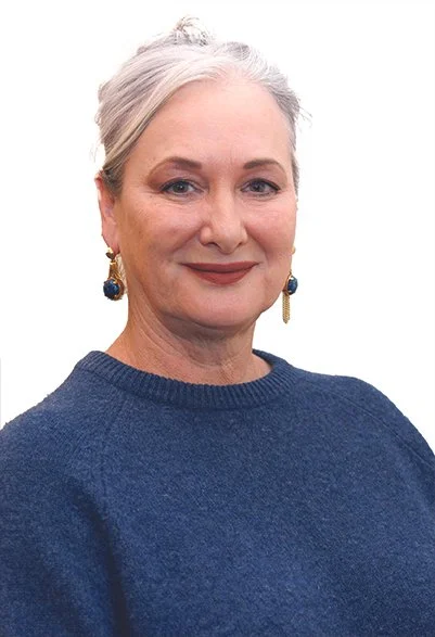 A woman with gray hair styled in an updo, wearing a dark blue sweater and gold earrings with blue stones, smiling at the camera against a plain white background.