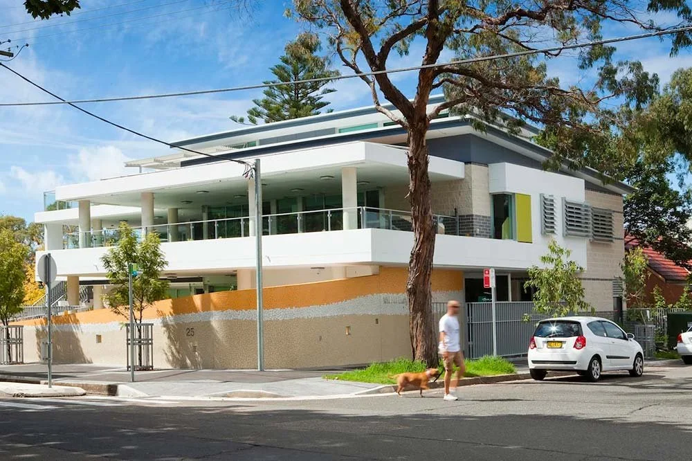 Modern two-story house with large balconies, glass railings, and a mix of white, gray, and orange exterior walls, on a sunny day with trees and a person walking a dog nearby.