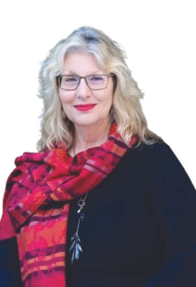 A middle-aged woman with blonde curly hair, wearing glasses, red lipstick, a red patterned scarf, and a black top.