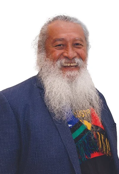 A smiling older man with a long white beard and white hair, wearing a dark blue jacket and a colorful T-shirt with abstract designs, against a white background.