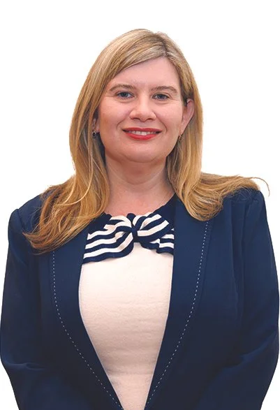 Portrait of a woman with blonde hair wearing a dark blazer and a striped bow tie blouse, smiling against a white background.