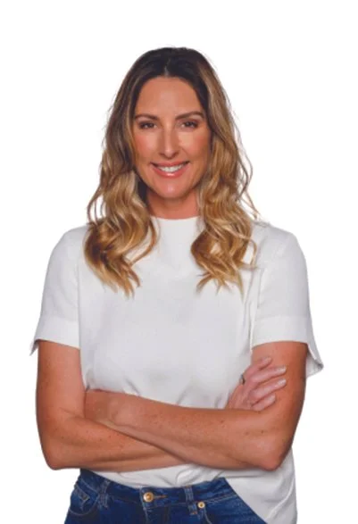A woman with wavy, shoulder-length blonde hair, smiling, wearing a white t-shirt and blue jeans, standing with arms crossed against a plain white background.