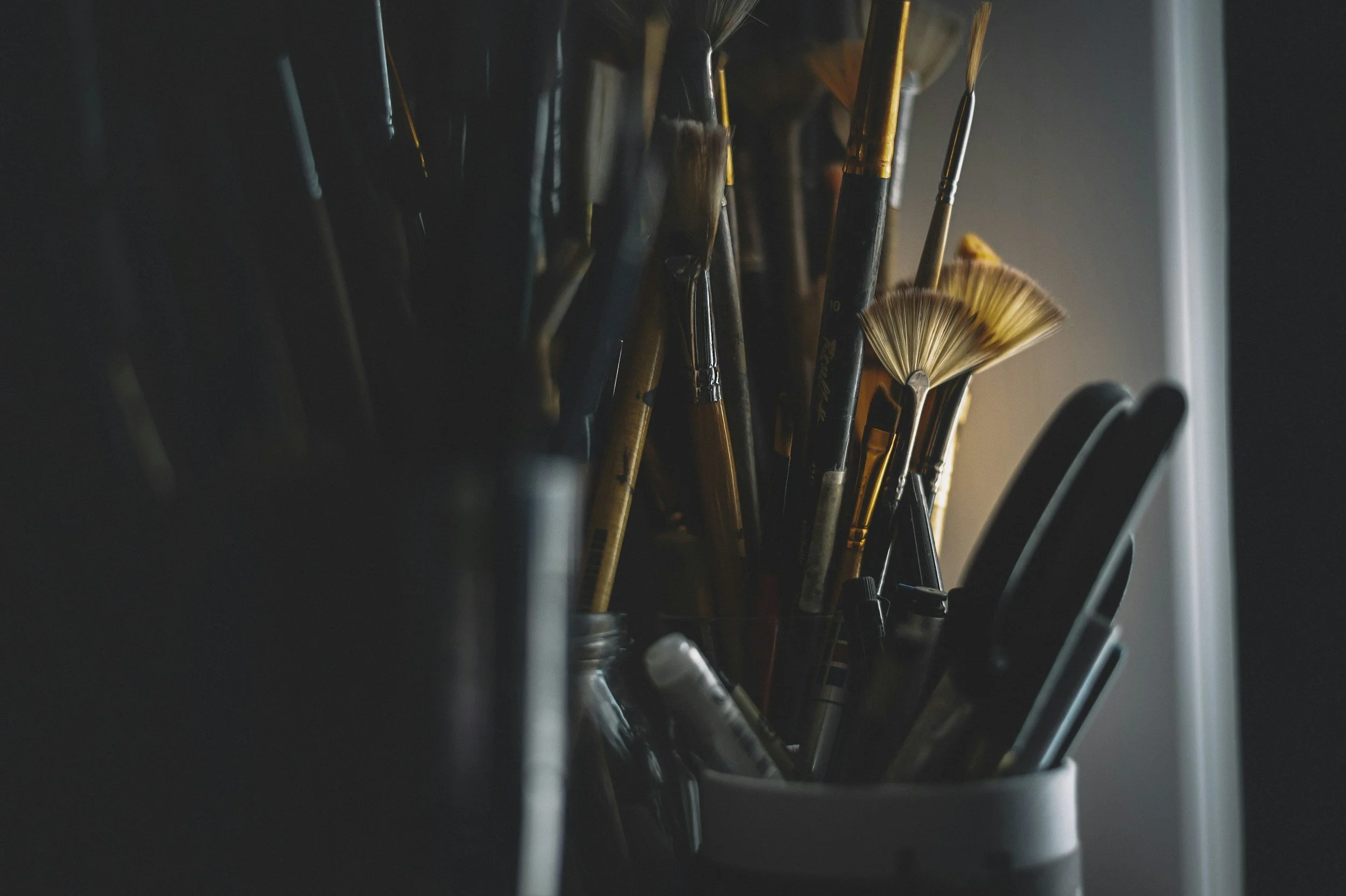 A collection of paintbrushes of various sizes in a container, with some brushes having bristles fanned out. The brushes are shown in a close-up shot in a dimly lit environment.