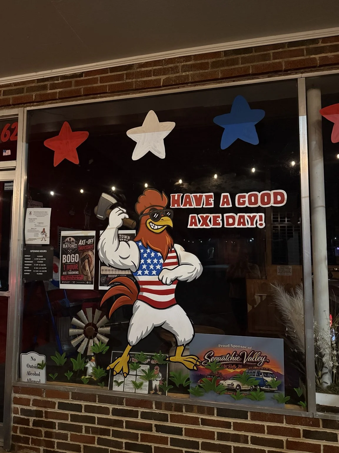 Hand-painted Fourth of July storefront window painting for a Tennessee axe throwing business (Small Town Hatchet) featuring a patriotic rooster, festive stars, and custom lettering by Blush Pony Studio.