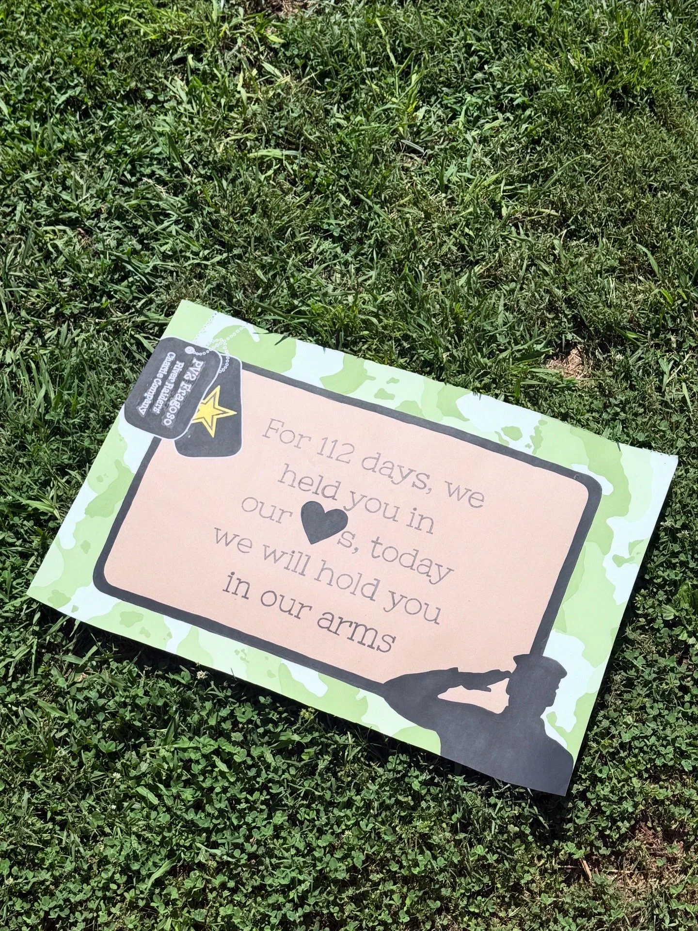 A memorial sign with a green camouflage border and silhouette of a soldier holding a rifle, placed on grass, with a message honoring a loved one.