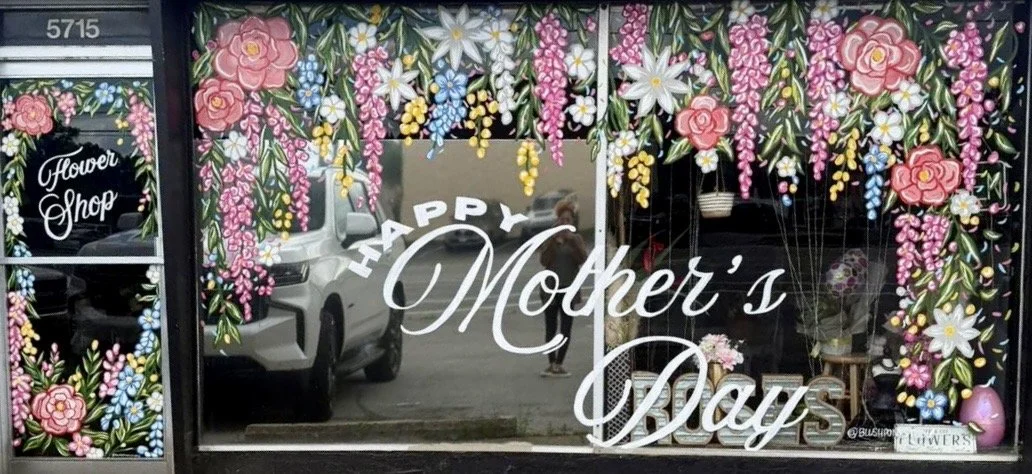 Shop window decorated with pink, white, yellow, and blue floral designs and a message for Mother's Day.