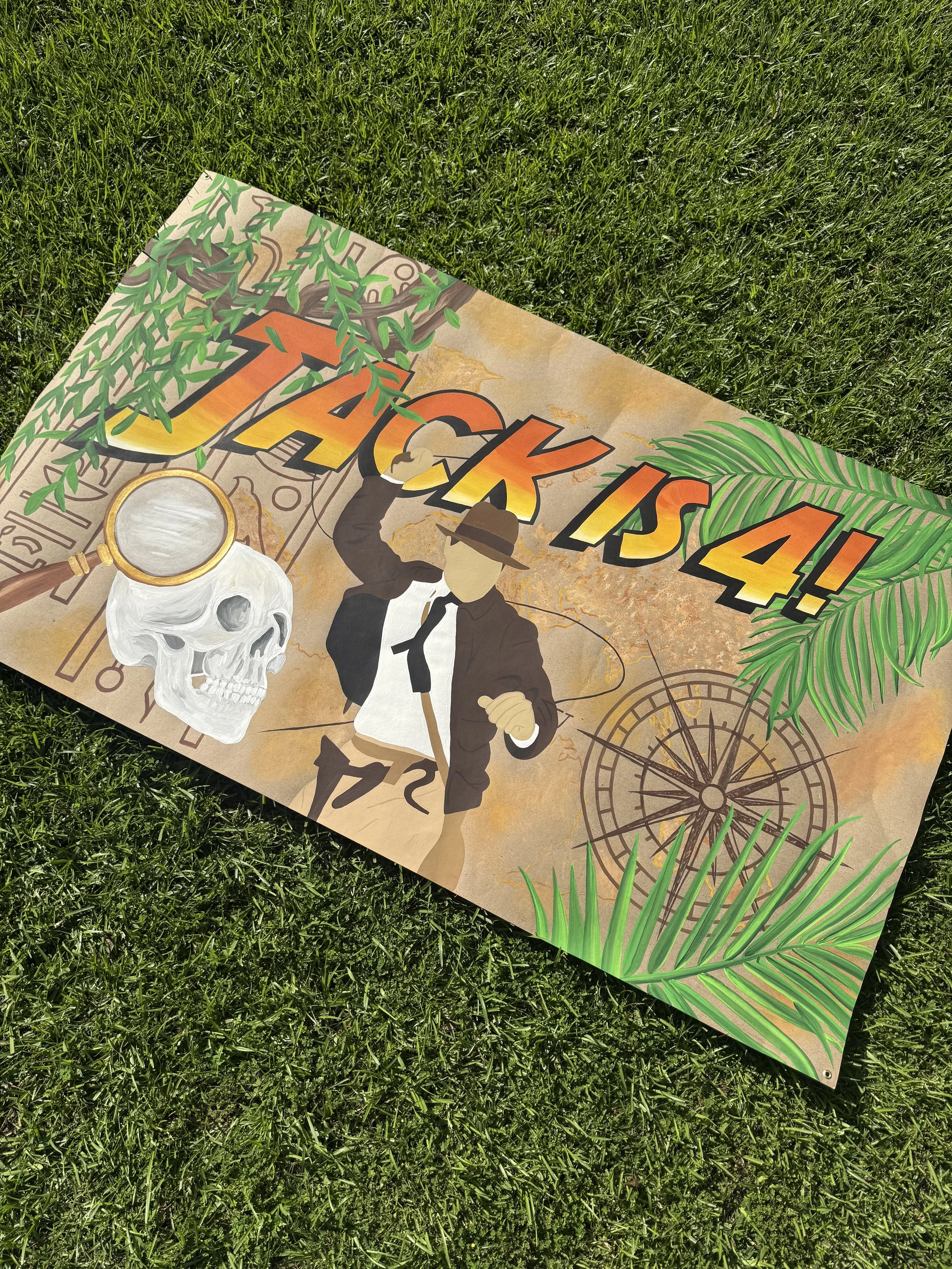 A colorful custom painted poster on green grass featuring a skull, a man in a suit and hat holding a magnifying glass, a large palm leaf, and the words 'JACK IS 4!'
