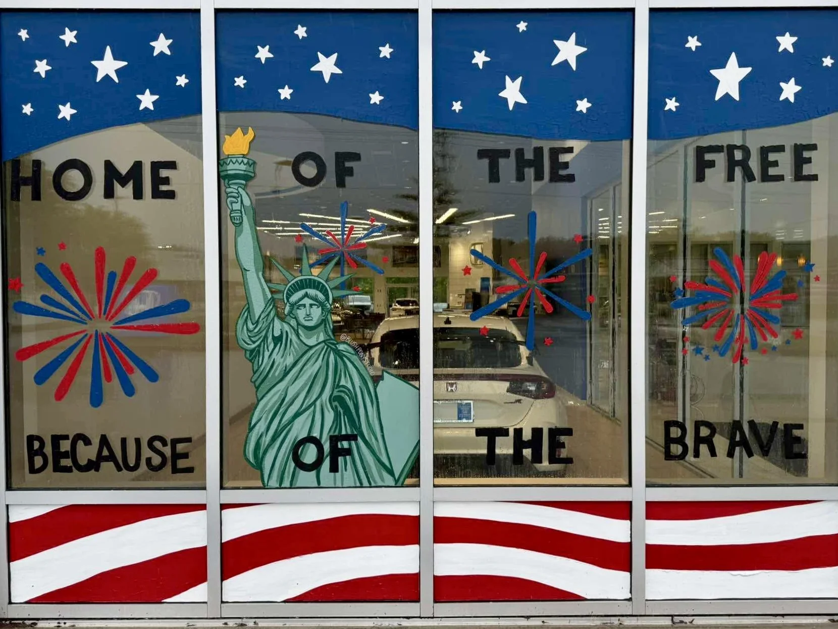 Decorated window with red, white, and blue fireworks and stars, featuring a drawing of the Statue of Liberty and the words "Home of the Free Because of the Brave."