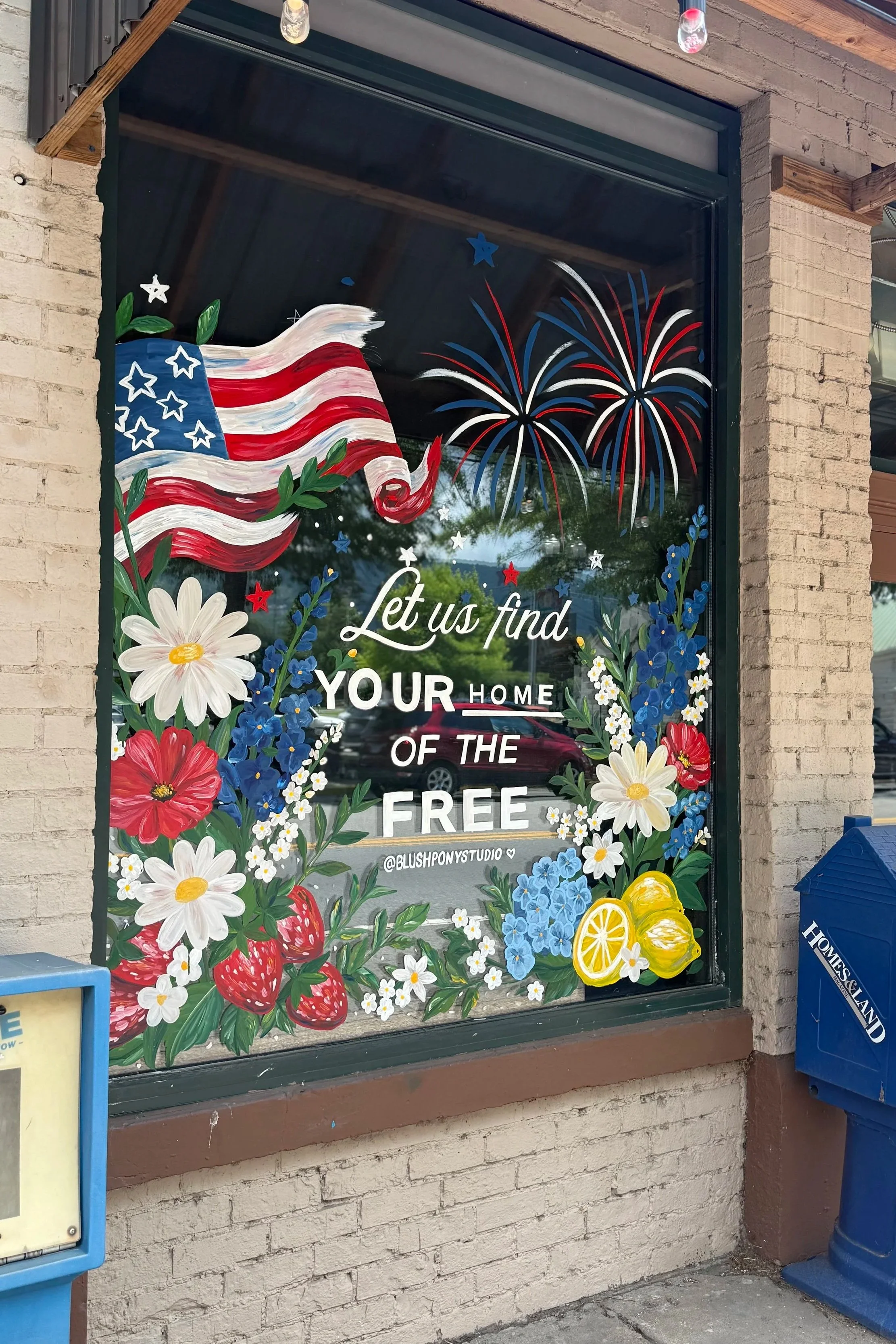 Decorative window display with a painted American flag, fireworks, and colorful flowers including daisies, strawberries, and lemons, along with the text 'Let us find your home of the free' and the Instagram handle '@blushponystudio'.