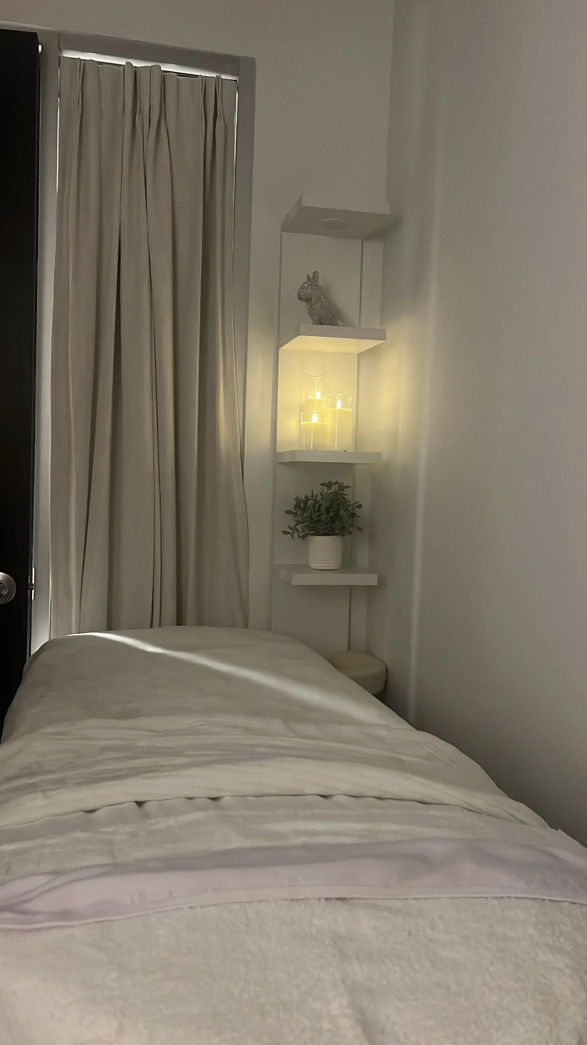 A cozy bedroom corner with a bed, beige bedding, and a white wall. There is a white wall-mounted shelf with a decorative bunny figurine, a small shelf with candles, and a potted green plant. Beige curtains cover a window.