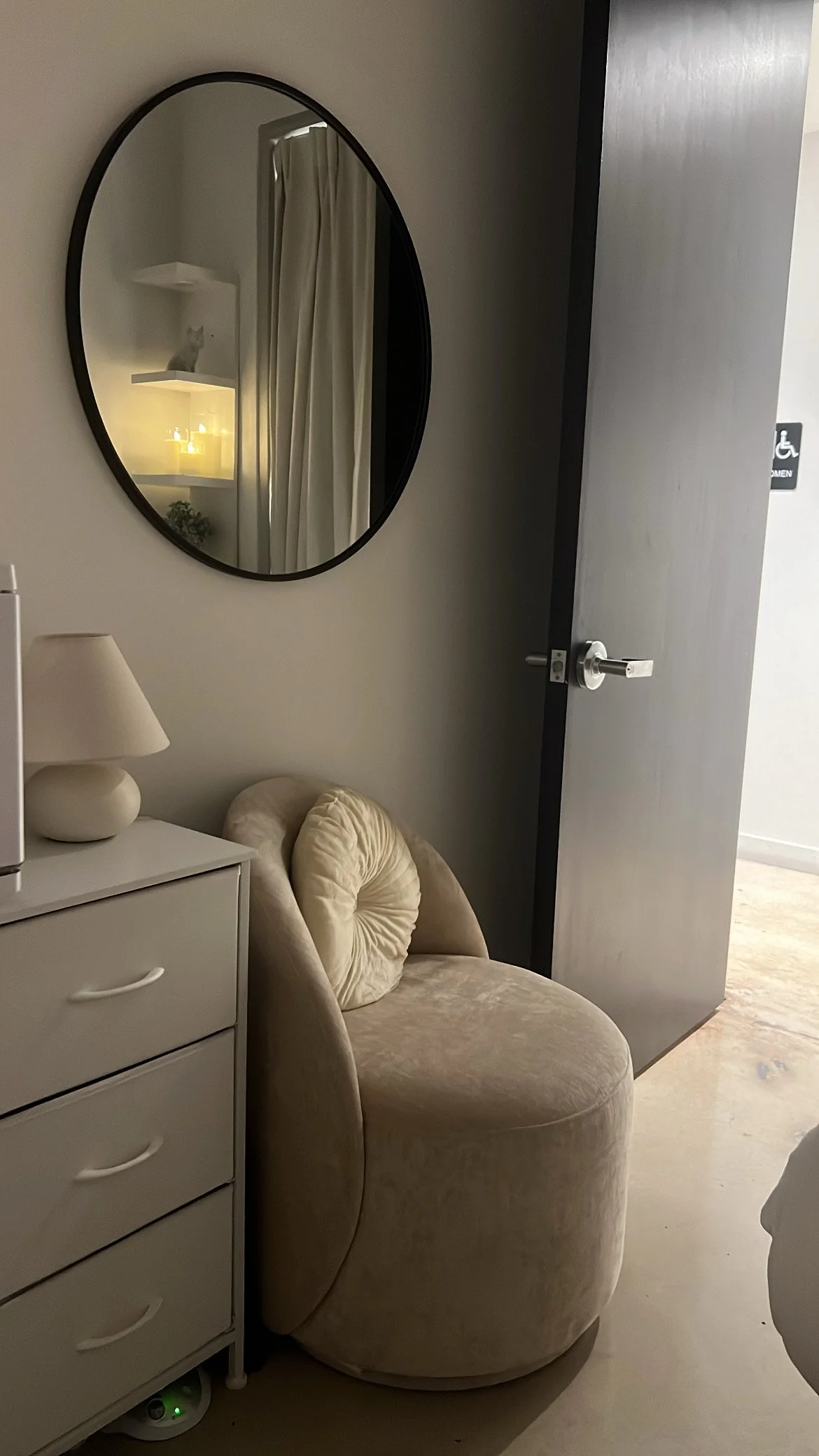 A cozy corner with a beige velvet armchair, a small white nightstand with a lamp, and a large round mirror on the wall reflecting a cat and candles. A curtain and door are also visible.